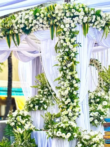A classic and elegant outdoor wedding mandap, constructed with a four-pillar structure, draped in sheer white fabric, and heavily adorned with fresh white flowers and greenery.