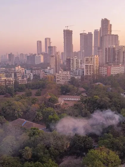 A drone shot captures smoke rising from a green, forested area in Mumbai, with the hazy city skyline in the background.
