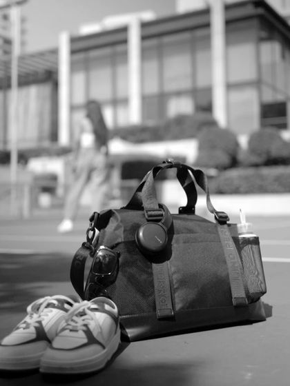 An atmospheric, black-and-white product shot for Lino Perros. Placing the gym bag, shoes, and accessories in the foreground with the model blurred in the back creates a lifestyle context while keeping the product as the hero.