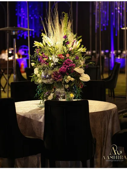An intimate dining table for a modern sangeet, featuring a dramatic centerpiece of wild lilies and purple florals, set against the cool blue glow of the event's lighting.
