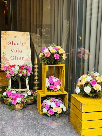 A vibrant and rustic welcome setup for a Haldi ceremony, creating the perfect 'Shadi Vala Ghar' feel. A custom sign is surrounded by colorful flower arrangements placed on bright yellow wooden crates.