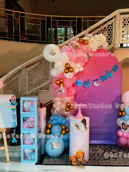 A grand baby shower setup by a staircase. The decor includes pink and blue balloons, butterfly accents, 'BABY' blocks, and a large 'Oh Baby' sign.
