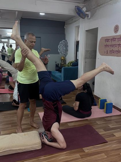 An instructor provides support to a student practicing a variation of Sirsasana (Headstand), ensuring stability and safety while exploring this advanced inversion.