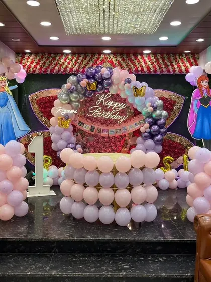 A princess-themed first birthday featuring characters from Frozen and Barbie. The stage is decorated with balloon pillars, character standees, and a balloon arch in pastel colors.
