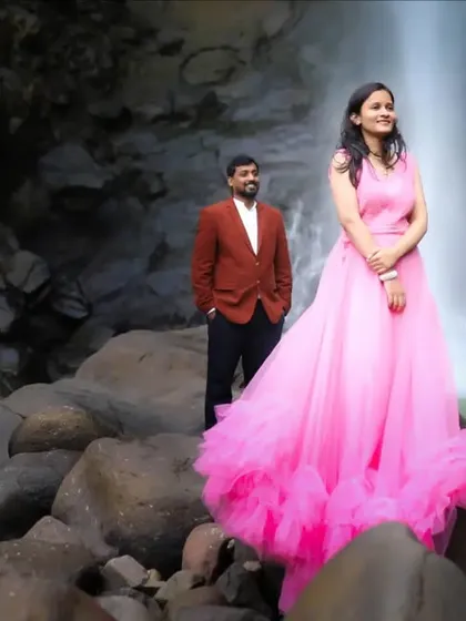 A stylish pre-wedding portrait featuring the bride in a vibrant pink gown, standing confidently in front of a beautiful waterfall, showcasing a blend of fashion and nature photography.