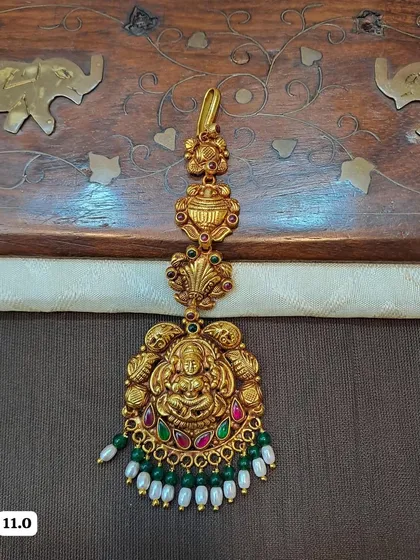 This maang tikka features a Goddess Lakshmi motif and is made from hallmarked 92.5 silver with gold plating. It is adorned with green and red stones, and finished with pearl and green bead drops.