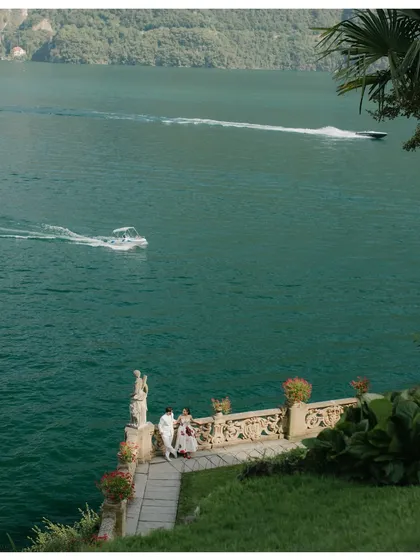A wide, scenic shot of a couple on a beautiful stone terrace overlooking the vibrant blue waters of Lake Como, Italy. The image captures the grandeur and serenity of the location.