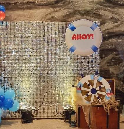 A shimmering photo backdrop for a nautical party, featuring a sequin wall, balloon garland, and a captain's wheel prop.