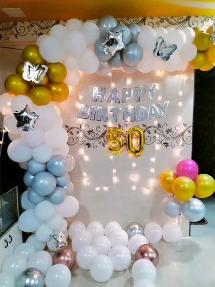 We offer simple yet beautiful birthday backdrops for home celebrations. This 50th birthday setup includes a balloon arch in white, silver, and gold, with butterfly accents and fairy lights.