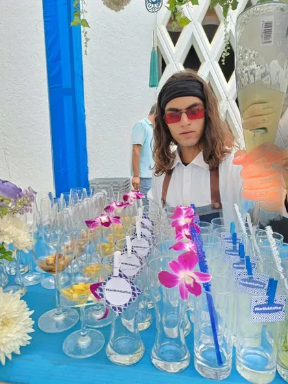 My bartender, dressed to match the theme, pours a drink for a guest. The bar is lined with glasses adorned with custom tags and purple orchids, showcasing the attention to detail in this Santorini-inspired setup.