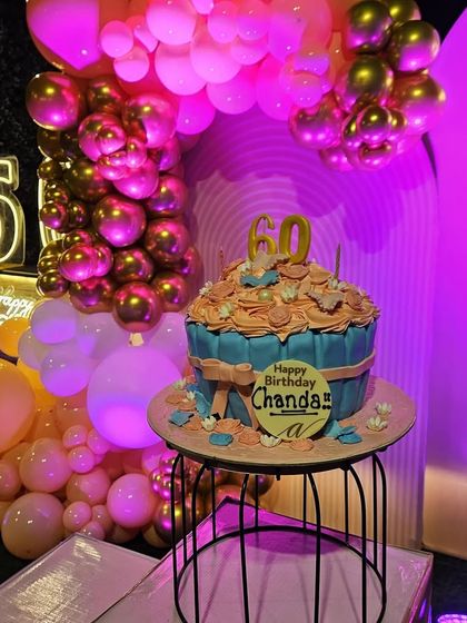 A close-up of a vibrant 60th birthday celebration cake display. The background features a pink arch, a 'Let's Party' neon sign, and a cascade of pink and chrome gold balloons under dramatic pink lighting.