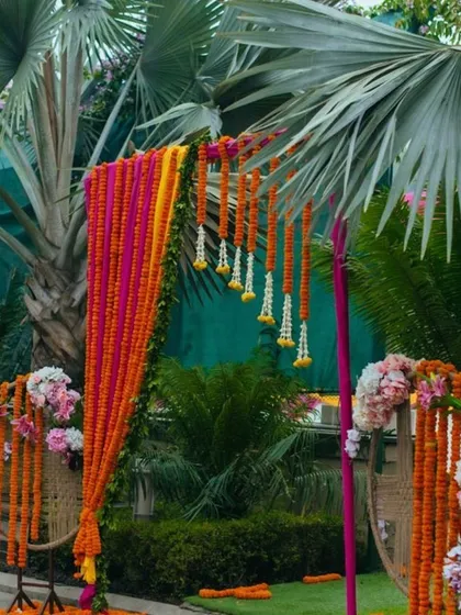 This is an outdoor entrance arch for a day event, set against lush greenery. The arch is decorated with strings of orange and pink marigolds, with floral arrangements on the side, creating a vibrant welcome.