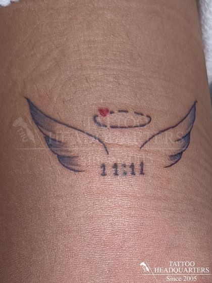 A small tattoo of angel wings with a halo and the numbers "11:11," symbolizing wishes and spiritual guidance.