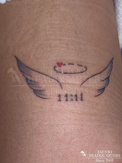 A small tattoo of angel wings with a halo and the numbers "11:11," symbolizing wishes and spiritual guidance.