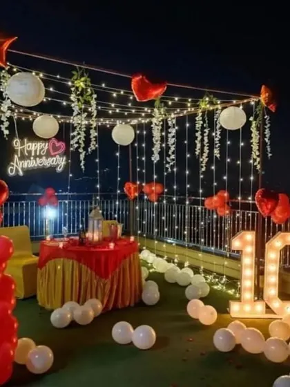 A romantic 10th-anniversary celebration on a rooftop. The decor features a canopy of fairy lights and paper lanterns, a 'Happy Anniversary' neon sign, and a light-up number '10'.