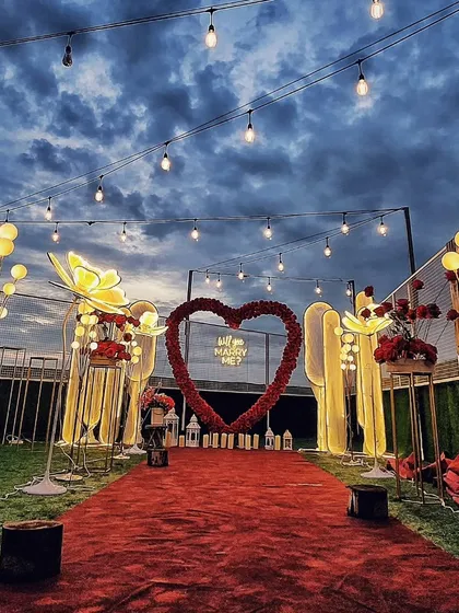 I design grand, dramatic proposal settings that make a statement. This setup features a long red carpet aisle lined with illuminated floral stands and candles, leading to a large red rose heart arch and a "Will you Marry Me?" sign.