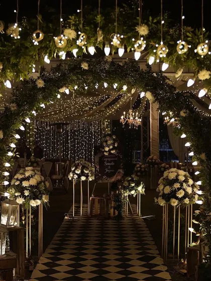 A glamorous Sangeet passage with an arch of greenery and white flowers, lit by hanging bulbs. The checkered floor and fairy light ceiling create a chic, party-ready atmosphere.