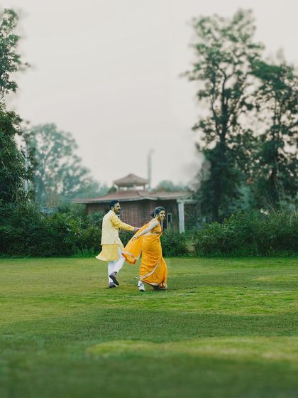 A couple shares a joyful run across a vast green lawn during their Haldi ceremony. The vibrant traditional outfits contrast beautifully with the natural setting, capturing the energy of the celebration.