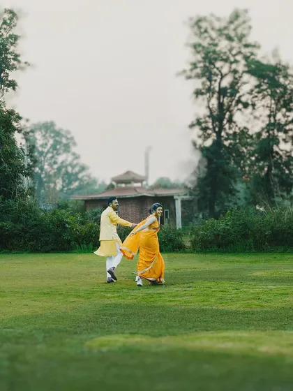 A couple shares a joyful run across a vast green lawn during their Haldi ceremony. The vibrant traditional outfits contrast beautifully with the natural setting, capturing the energy of the celebration.
