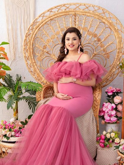 Seated on a grand peacock chair, this mother-to-be looks like royalty in a vibrant pink mermaid gown. The surrounding floral arrangements in matching tones create a lush, romantic scene.