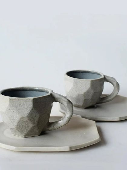 A modern, geometric cup and saucer set with a faceted design. The matte grey finish gives it a contemporary and sophisticated look.