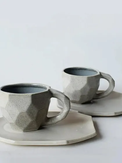 A modern, geometric cup and saucer set with a faceted design. The matte grey finish gives it a contemporary and sophisticated look.