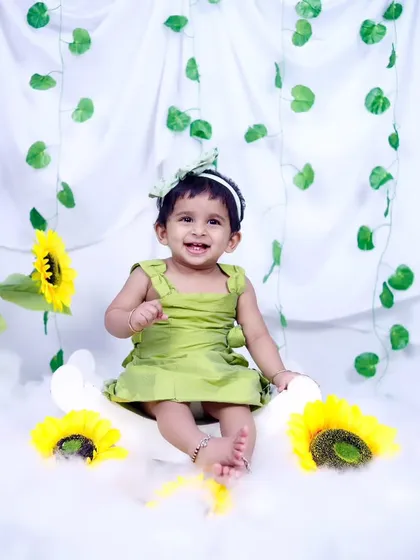 A happy baby girl in a green dress, sitting among sunflowers and clouds. Her bright smile lights up the photo.
