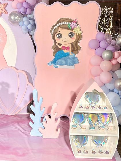 A closer look at the details from a mermaid-themed birthday party. This setup includes a boat-shaped shelf for party favors and a cute mermaid cutout against a pink backdrop, perfect for an under-the-sea adventure.