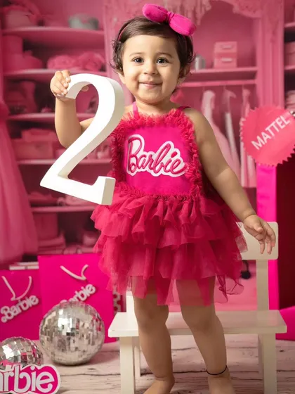 Celebrating turning two with a Barbie theme. This little girl proudly holds up the number '2' in her pink dress, surrounded by disco balls and Barbie accessories.