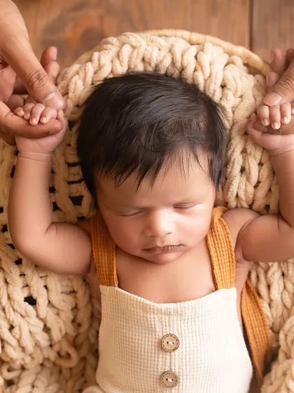 A newborn sleeps peacefully in a woven basket, his tiny hands held by his parents. This simple, rustic setup highlights the baby's delicate features and the protective love surrounding him.