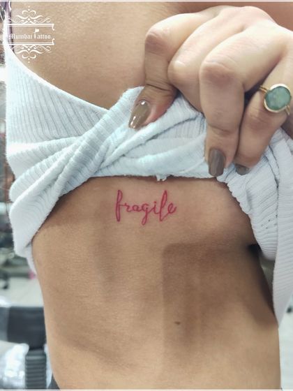 The word "fragile" in red ink on the ribcage. Using red ink for script tattoos creates a soft yet striking effect, and we ensure the fine lines are executed perfectly.
