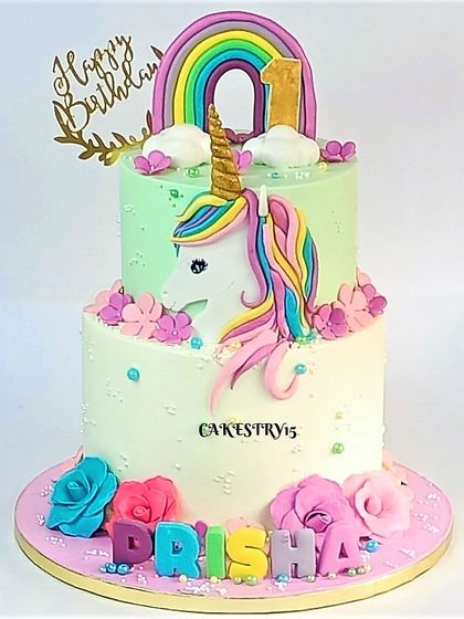 A lovely two-tier unicorn cake for a first birthday. This design features a large fondant unicorn head against a soft green tier, with a rainbow and colorful roses below.