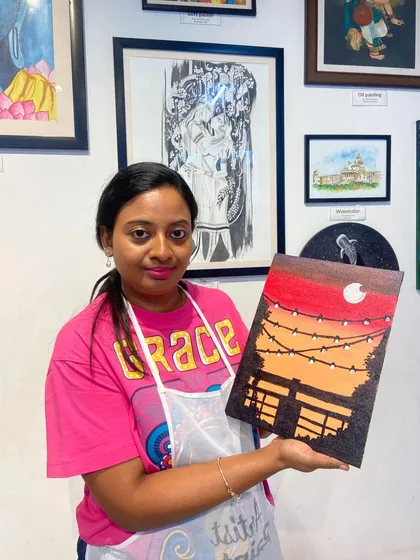 A participant with her finished canvas painting of a sunset and string lights. It's a popular theme that creates a warm and cozy atmosphere.