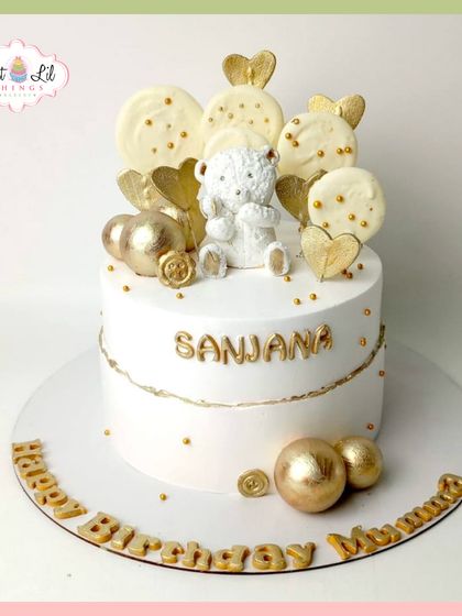 Pure elegance in gold and white. This dreamy birthday cake is topped with a white chocolate teddy bear, lollipops, and golden spheres.