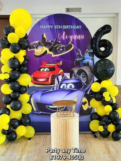 A Batman and Hot Wheels theme for Avyaan's 6th birthday. The decoration includes a custom printed backdrop with cars, black and yellow balloon columns, and a large number 6 foil balloon.