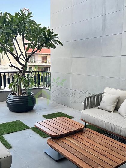 A balcony in Yojna Vihar, Delhi, designed for relaxation. The space features modern woven furniture, a large potted tree, and patches of artificial grass set among the tiles for a touch of green.