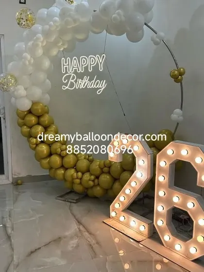 A sophisticated gold and white theme for a 26th birthday. The half-moon balloon arch is paired with a neon sign and large, brightly lit marquee numbers.