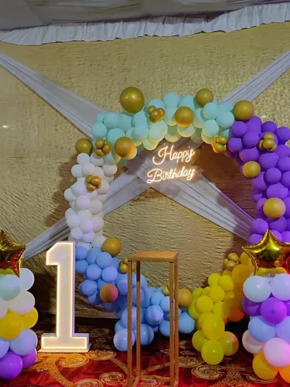 A colorful and bright setup for a first birthday party. This decoration includes a multi-colored balloon ring, a light-up number '1', and balloon pillars topped with star foil balloons.