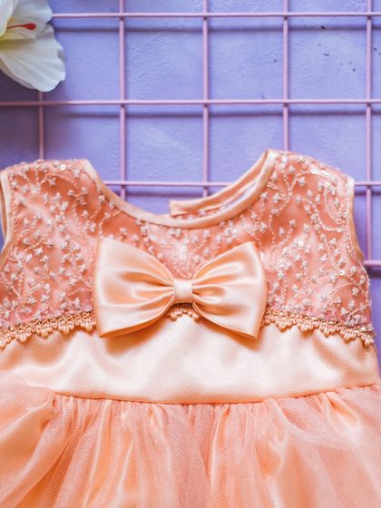 This peach-colored dress features a bodice with intricate floral embroidery and a delicate lace trim. A satin bow completes this sweet and sophisticated design.