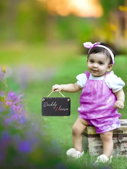 An adorable outdoor shot of a baby girl in a pink outfit, sitting on a trunk and holding a small sign with my brand name. The lush green and purple flowers in the background add a beautiful touch.