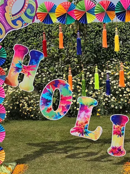 A vibrant and fun photo booth setup for a Holi party, featuring a colourful arch, paper fans, and 'Holi' letters.