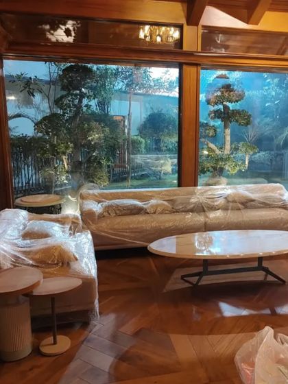 The living area within the gazebo, with furniture still protected, looks out onto the lush garden through floor-to-ceiling glass walls, perfectly blending the interior with the natural world outside.