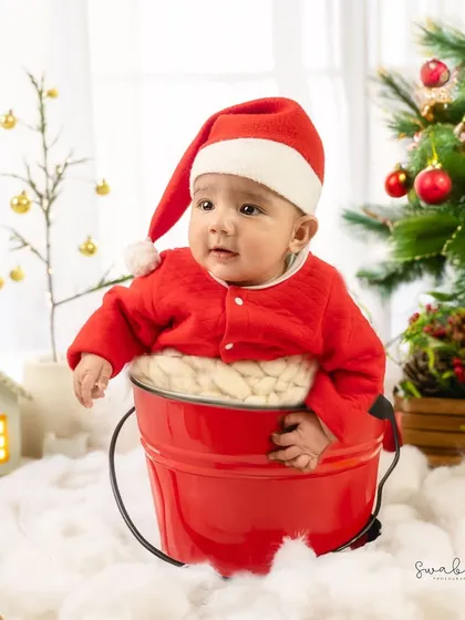 Popping out of a bright red bucket, this baby Santa looks curious and adorable next to a beautifully decorated Christmas tree.