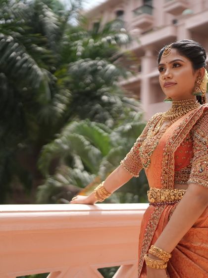 The bride stands on a balcony, her magnificent orange and gold saree catching the light. This full length shot shows the grandeur and elegance of a traditional bridal outfit designed for a palace wedding feel.