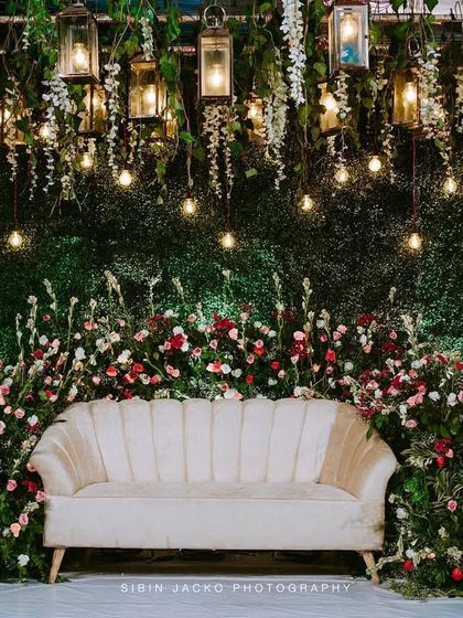A romantic stage setup with a lush green wall, a cream-colored sofa, and an overhead installation of hanging lanterns and Edison bulbs.