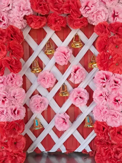 A compact backdrop design featuring a frame of red and pink artificial carnations. The background uses a white lattice pattern over red fabric, decorated with hanging golden bells and pom-poms.