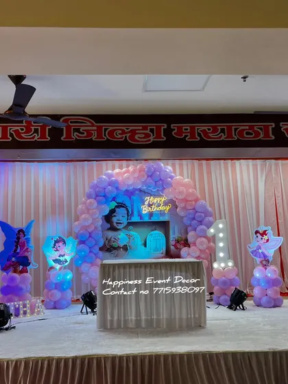 A full stage setup for a 'Pari' or fairy-themed first birthday in a community hall. The decor includes a balloon arch, a custom photo backdrop, and multiple fairy cutouts.