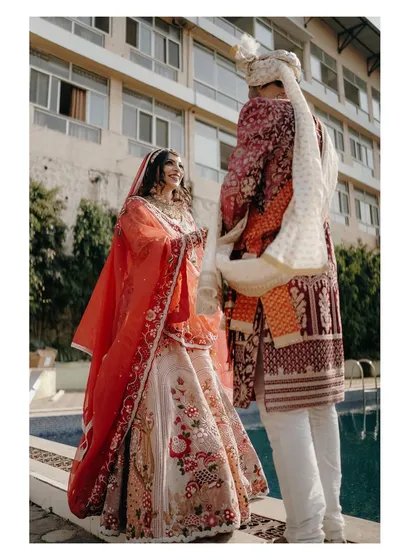 A candid moment by the poolside. The bride looks at her groom with a beautiful smile, her vibrant red and white lehenga creating a stunning visual against the modern backdrop.