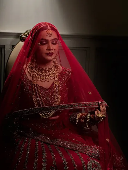 A candid moment under the veil. The soft lighting highlights the flawless base and the deep red tones of the makeup, creating a dreamy, romantic bridal portrait.