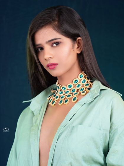 A beauty-focused portrait highlighting a statement necklace. The simple styling with an unbuttoned shirt draws all attention to the jewelry and the model's expression.